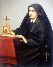 A nun in black robes sits at a table writing, with a small religious icon and a cross nearby. The serene presence of Anna Rosa Gattorno, now blessed, seems to inspire her quiet devotion.