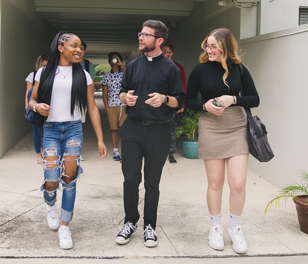 A priest walks and chats with two young women in casual attire, sharing smiles and heartfelt testimonials outdoors.