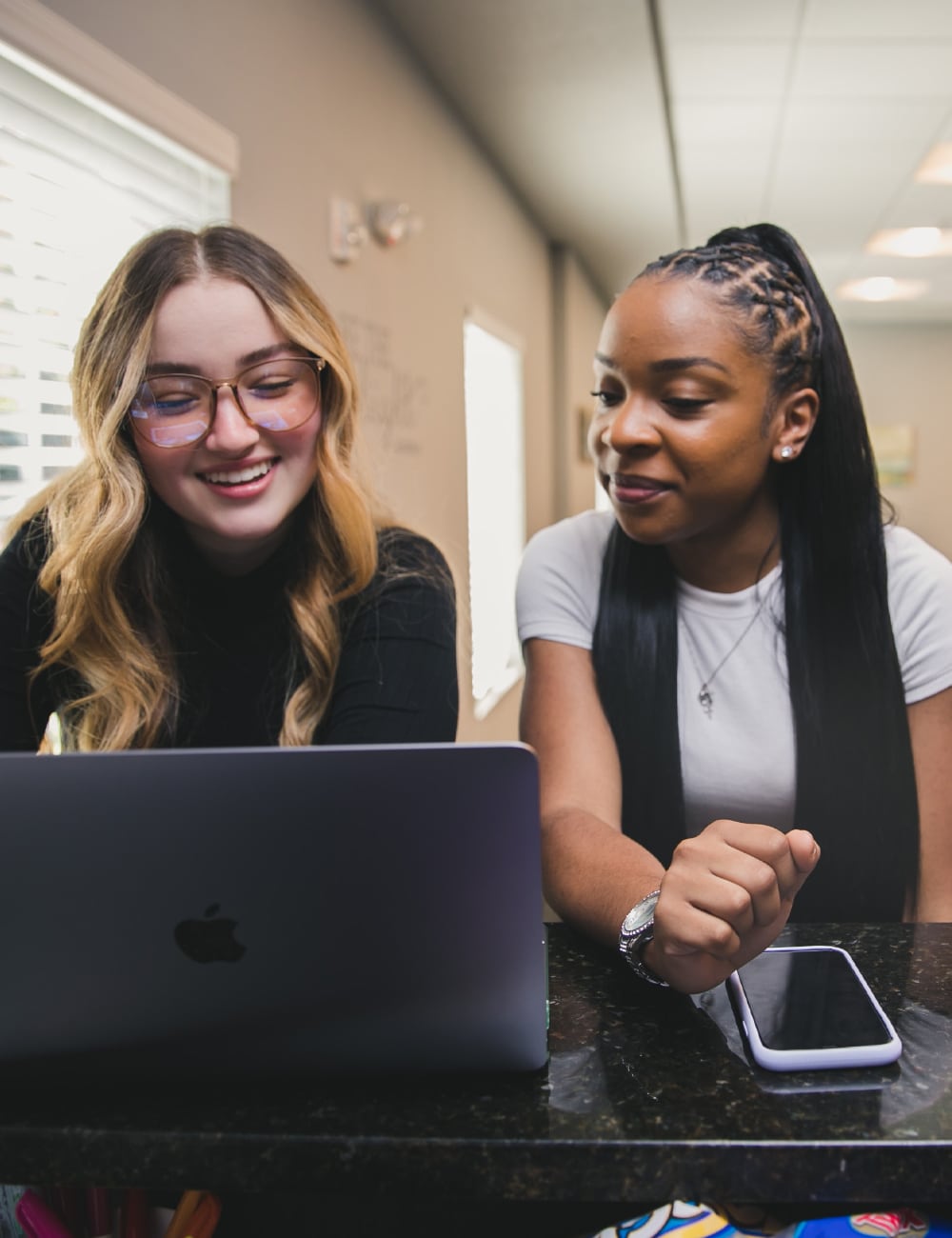 Two women smiling while looking at a laptop in a cozy office setting, reminiscent of a home workspace. A smartphone is conveniently placed on the table beside them.