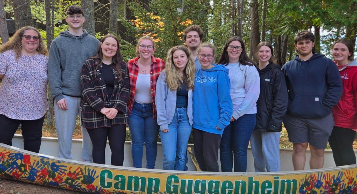 A group of people stands together outdoors behind a sign reading Camp Guggenheim, surrounded by trees, ready to share their testimonials.