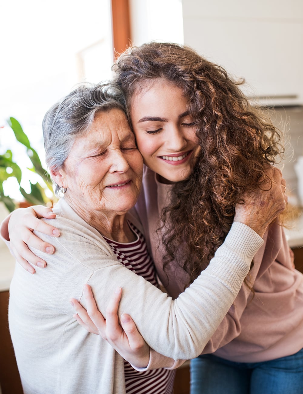 Young woman with curly hair hugging an elderly woman, both smiling warmly in a cozy room.